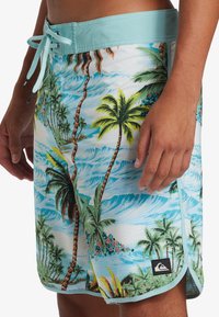 Men's swim shorts in a light blue waistband with a tropical palm tree print in greens and yellows on a white background; side slit.