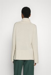 En person vänd bort från kameran, som bär en krämfärgad ribbad polotröja och mörkgröna byxor, mot en enkel ljus bakgrund.