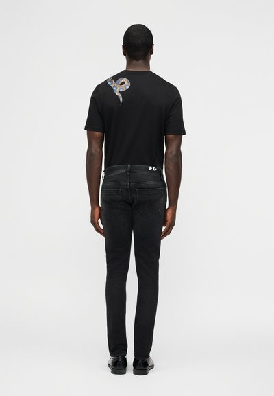 Homme portant un jean noir et un t-shirt noir avec un graphique de serpent bleu et beige sur le haut du dos, debout devant un fond blanc uni.