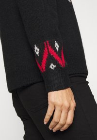 Schwarzer gestrickter Pullover mit rot-weißer geometrischer Musterung an den Bündchen. Weiche Textur und lässige Passform, kombiniert mit schwarzen Hosen.