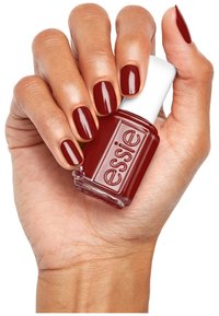Vernis à ongles rouge foncé dans un flacon en verre étiqueté "essie", tenu par une main avec des ongles soignés peints de la même teinte. Fini lisse, aspect brillant.