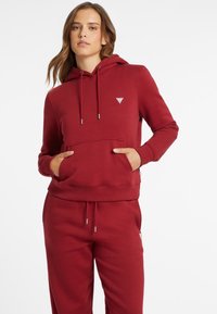 Vrouw in een rode hooded sweatshirt en bijpassende joggingbroek, staand met handen in de zakken van het sweatshirt tegen een witte achtergrond.