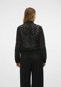 Blouson en dentelle noire avec motifs floraux, poignets côtelés et silhouette courte, porté sur un pantalon noir.
