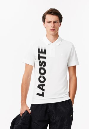 Chemise polo blanche avec logo "LACOSTE" noir, manches courtes et col classique. Portée avec un pantalon noir et tenant une veste noire.