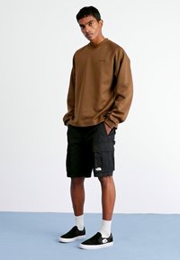 Hnědá dlouhá mikina s rukávy, černé cargo šortky s kapsami, černé slip-on boty a bílé ponožky do půli lýtek, na světlém pozadí.