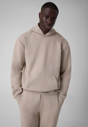 Young man wearing a beige hoodie and matching sweatpants, standing with hands in pockets against a plain light gray background.