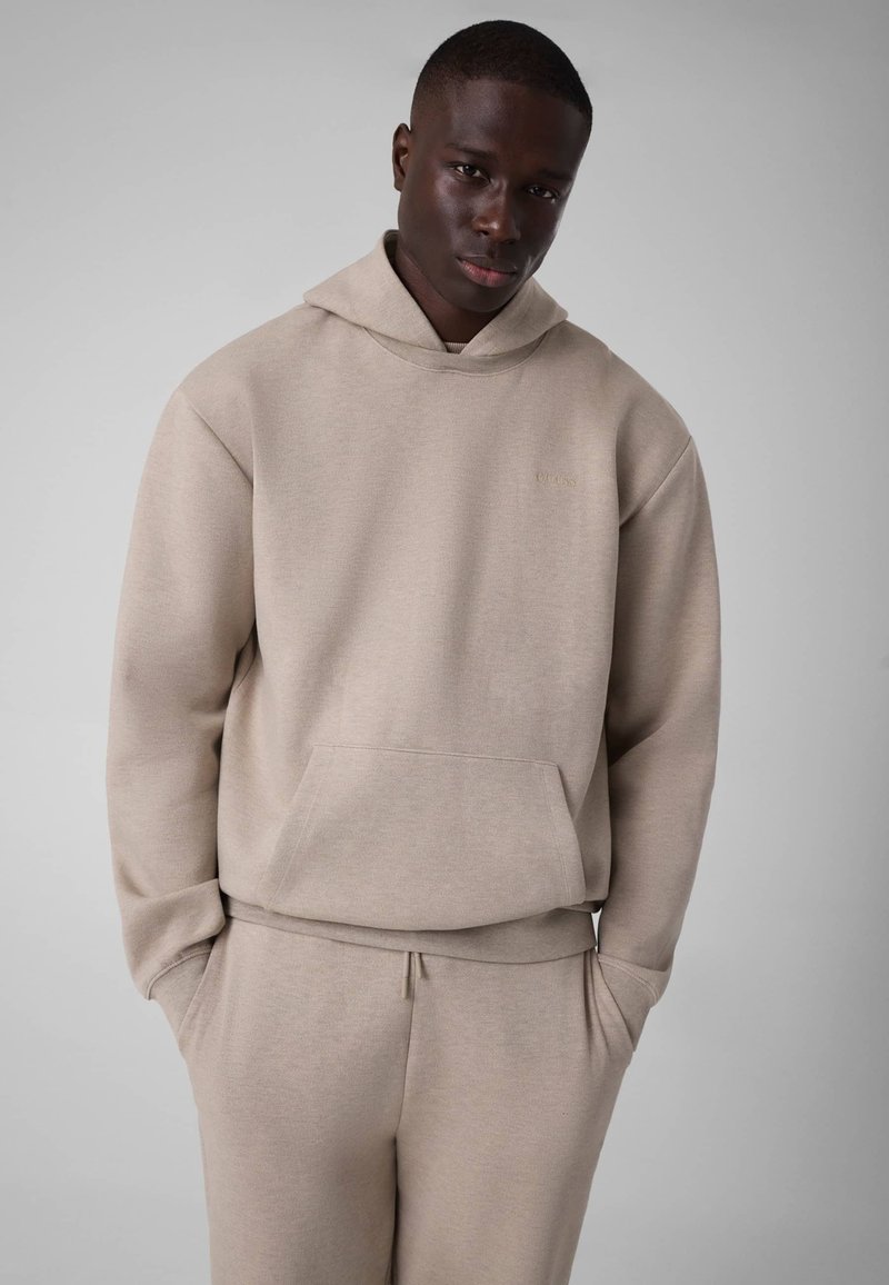 Jeune homme portant un sweat à capuche beige et un pantalon de survêtement assorti, debout les mains dans les poches sur un fond gris clair uni.