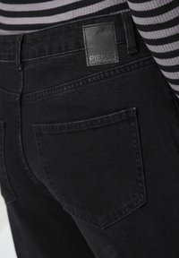 Μαύρα τζιν από ντένιμ με ετικέτα παPatch που αναγράφει "PIECES", πίσω τσέπες και λεία, ματ υφή. Λωρίδα μπλούζα μερικώς ορατή.