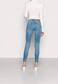 Mujer vistiendo jeans skinny de color azul claro, un suéter de cuello alto blanco con canalé y zapatillas blancas, de pie con la espalda hacia la cámara sobre un fondo liso.