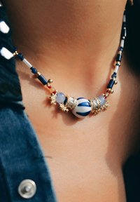 Le collier en perles présente un centre en rayures bleues et blanches, des accents dorés, et un mélange de petites perles en bleu, rouge et blanc.