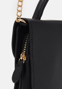 Mustan tekonahkaisen käsilaukun, jossa on kullanväriset yksityiskohdat, vetoketjukiinnitys, ketjuhihna ja tyylikäs muotoilu.