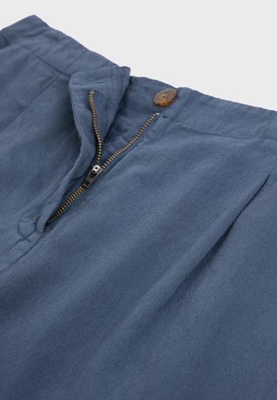 Niebieskie sztruksowe spodnie z otwartym mosiężnym zamkiem i brązowym guzikiem w pasie na białym tle.