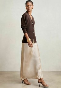 Cárdigan marrón de punto con cuello en V y grandes botones, combinado con una falda maxi de satén crema y sandalias de tacón abierto en nude. Acentos de pulsera dorada.
