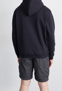 Felpa nera con cappuccio e polsini a coste, abbinata a pantaloni cargo grigi con tasche laterali. Cuciture visibili e texture morbida.