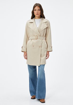 Trench coat beige con bottoni doppi, ampie alette e cintura, abbinato a jeans chiari a zampa d'elefante e stivali marroni a punta quadrata.