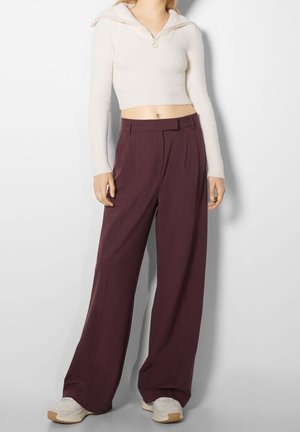 Avskuren krämfärgad tröja med dragkedja vid halslinningen, matchad med löst sittande plommonfärgade byxor. Vita sneakers fulländar outfiten. Slät textur.