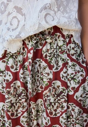 Weißes Spitzentop mit floraler Stickerei, kombiniert mit einem hoch taillierten Rock mit einem roten, weißen und grünen floralen geometrischen Muster, zusammengebunden mit einem Stoffgürtel.