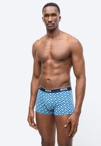 Μυώδης άνδρας στέκεται φορώντας μπλε μπόξερ με σχέδιο και μαύρη μέση με την ετικέτα "BOSS" σε απλό λευκό φόντο.