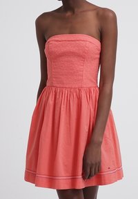 Korálové šaty bez ramínek s texturovaným živůtkem, rozšířenou sukní a vodorovnými pruhy na lemu. Vyrobené z lehkého bavlněného materiálu.
