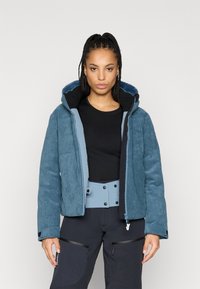 Modrá kordurovým sako s černou fleecovou kapucí, s uzávěrem na zip a bočními kapsami. Nosená přes černý top a modrý opasek.