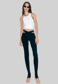 Svarta leggings med utskurna detaljer vid midjan, tillsammans med en vit ärmlös topp. Modellen bär solglasögon och platta sandaler. Slät textur.