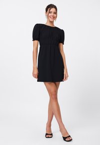 Robe noire courte avec manches bouffantes, taille froncée et texture lisse. Associée à des talons ouverts noirs. Design simple et épuré.