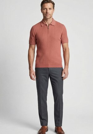 Man staat en draagt een roestkleurige, korte mouw geribbelde polo, donkergrijze broek en bruine leren loafers tegen een effen achtergrond.