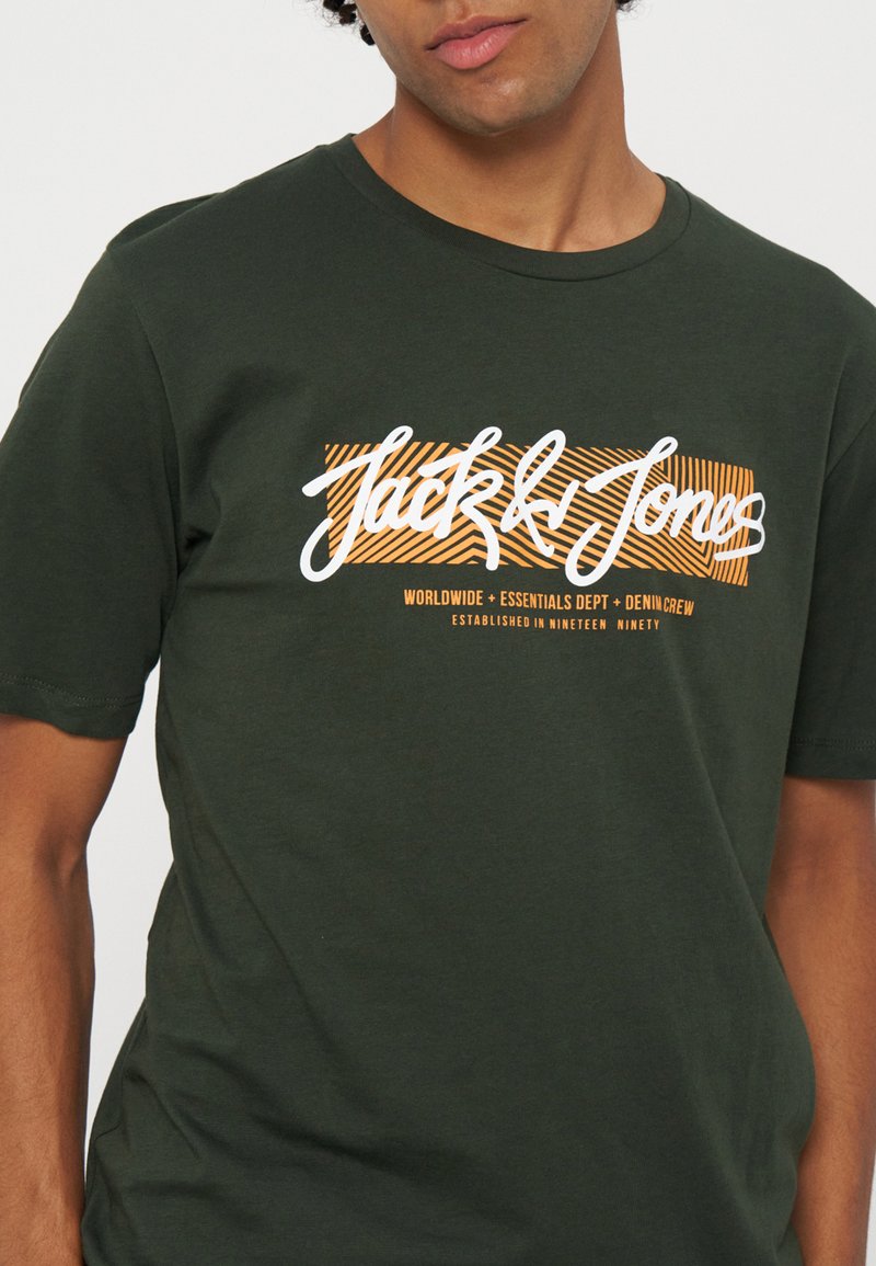 Hombre con camiseta verde oscuro con logo "Jack & Jones" en naranja y blanco y texto pequeño debajo en la parte delantera.