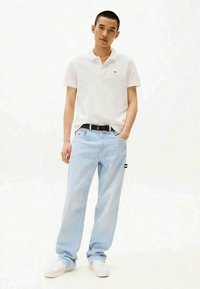 Maglietta polo bianca, pantaloni a righe blu chiaro e sneakers bianche. I pantaloni hanno una vestibilità rilassata, con un discreto patch logo sul lato.
