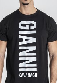 Čierne tričko z bavlny s veľkým bielym vertikálnym potlačeným textom, ktorý znie "GIANNI KAVANAGH" v tučnom, bezserifovom písmene.