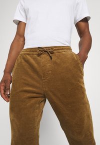 Bruine ribcord broek met verticale ribbels, elastische tailleband en trekkoord, weergegeven met een wit T-shirt. Voorkanten van de zakken zichtbaar.