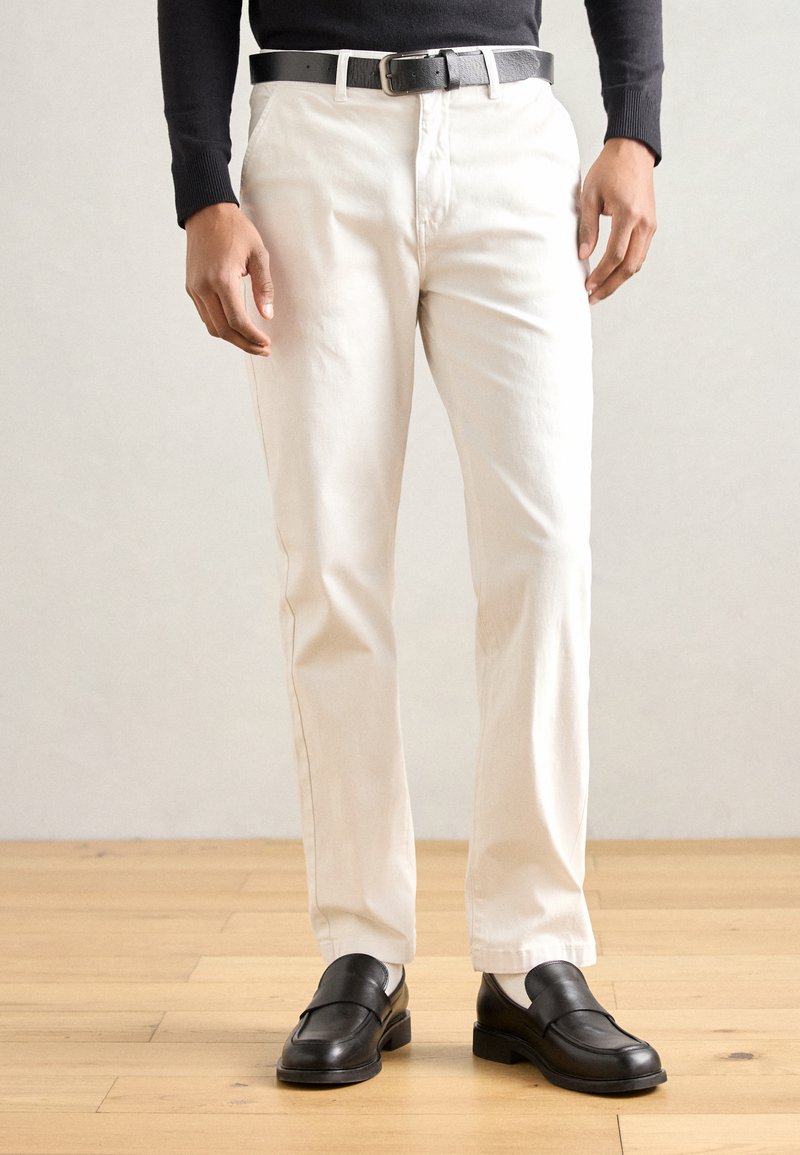 Białe bawełniane spodnie o prostym kroju, zestawione z czarnymi skórzanymi mokasynami i czarnym paskiem, noszone z dopasowaną czarną bluzką z długim rękawem.