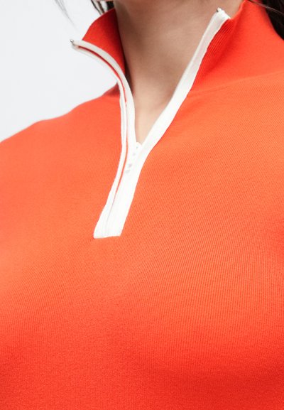 Pull-over à fermeture éclair quart de zip en tissu orange vif, avec fermeture éclair blanche et encolure assortie. Texture lisse et côtelée visible sur le vêtement.