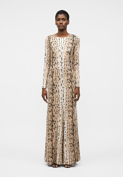 Femme debout portant une longue robe imprimée animal beige et noire à manches longues et des chaussures noires ouvertes sur un fond uni.