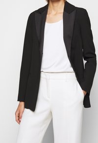 Blazer noir avec revers en satin, coupe ajustée et deux poches avant, porté sur un haut blanc et associé à un pantalon blanc ajusté.