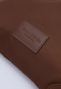 Hnědý textilní sáček s koženým štítkem s razeným názvem značky. Hladký povrch a obdélníkový tvar. Šité detaily na okrajích.