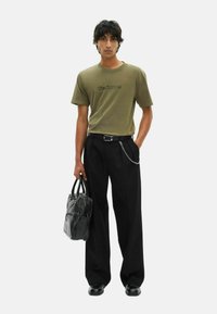 Joven con camiseta verde oliva y pantalones de pierna ancha negros, sosteniendo un bolso de cuero negro en una mano, de pie frente a un fondo liso.