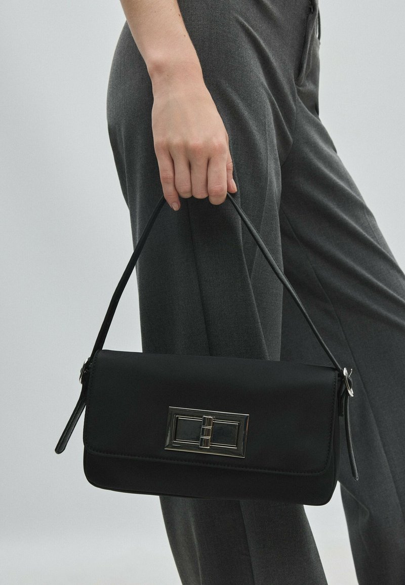 Main tenant un sac à main noir avec une fermeture rectangulaire argentée, sur un fond de pantalon gris foncé.