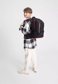 Zaino nero con accenti rossi, diverse tasche e spallacci regolabili, indossato da un bambino con una camicia a scacchi e pantaloni color crema.
