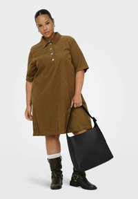 Robe en velours côtelé marron avec col et manches courtes, présentant des détails de boutons. Le mannequin porte un sac fourre-tout noir et des bottines noires à boucles.
