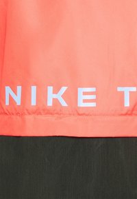 Nike-juoksutakki, jossa kirkkaan korallinen yläosa teksturoidulla kankaalla ja kontrastina tumma alaosa, jossa rohkea valkoinen brändäys.
