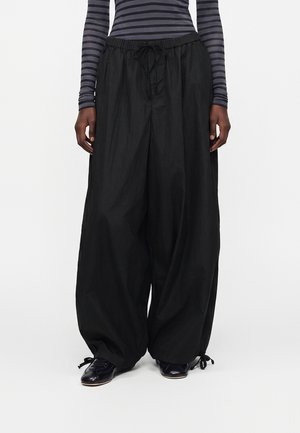 Persoană purtând pantaloni negri largi cu șnur și pantofi negri, asortați cu un top cu mâneci lungi cu dungi negre și gri, stând pe un fundal alb.