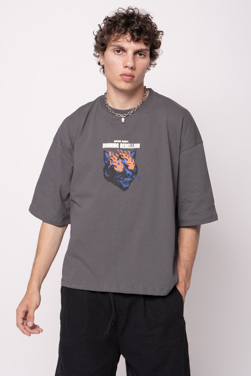 Šedé oversized tričko s krátkým rukávem, na kterém se nachází modrooranžový grafický motiv kočičí tváře obklopené plameny. Černé šortky doplňují tento outfit.