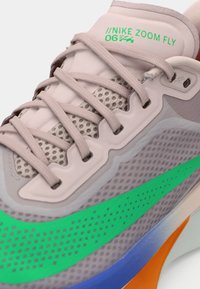 Gros plan de la chaussure Nike Zoom Fly avec des lacets beige, une tige en mesh gris, un swoosh vert vif, et des accents orange et bleu sur la semelle.