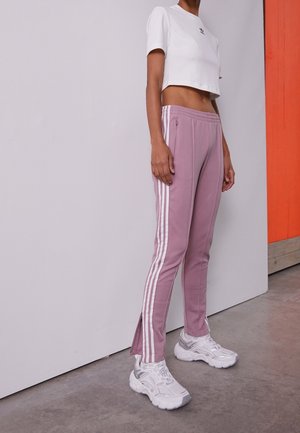 Person iført lilla træningsbukser med hvide striber, hvid cropped t-shirt og hvide chunky sneakers står ved en hvid og orange væg.
