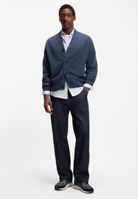 Mörkblå stickad kofta, knäppt, bärs över en vit skjorta; kombinerad med mörka denimbyxor och grå sneakers med texturerad yta.