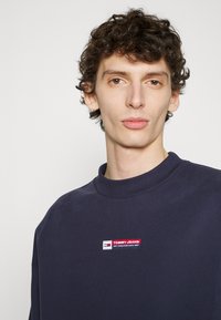 Tengerészeti pulóver, kerek nyakkivágással, bordázott mandzsettákkal és hímzett "TOMMY JEANS" logóval, piros, fehér és kék részletekkel a mellkason.