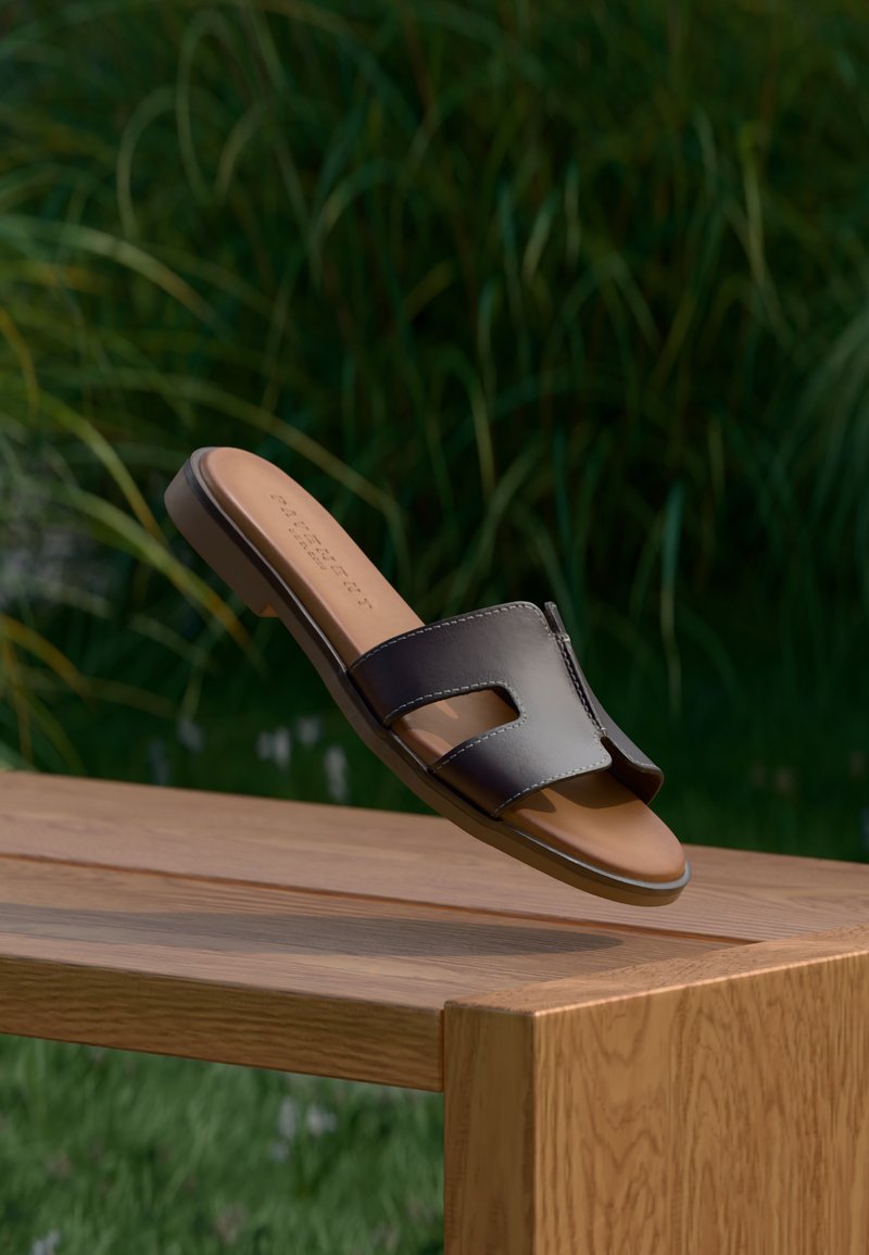 Sandale noire en cuir à enfiler, avec bout ouvert et double bride, dotée d'une semelle intérieure beige et d'une semelle plate, posée sur un fond d'herbe.