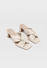 Des mules à talons carrés beige présentent un dessus en textile noué avec une finition lisse, un bout carré et un petit talon solide pour le confort et la stabilité.