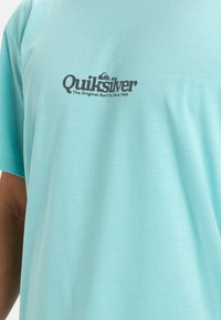 Camiseta de algodón de color azul claro con cuello redondo y mangas cortas; presenta un logo oscuro y texto en el pecho que indica el nombre de la marca.